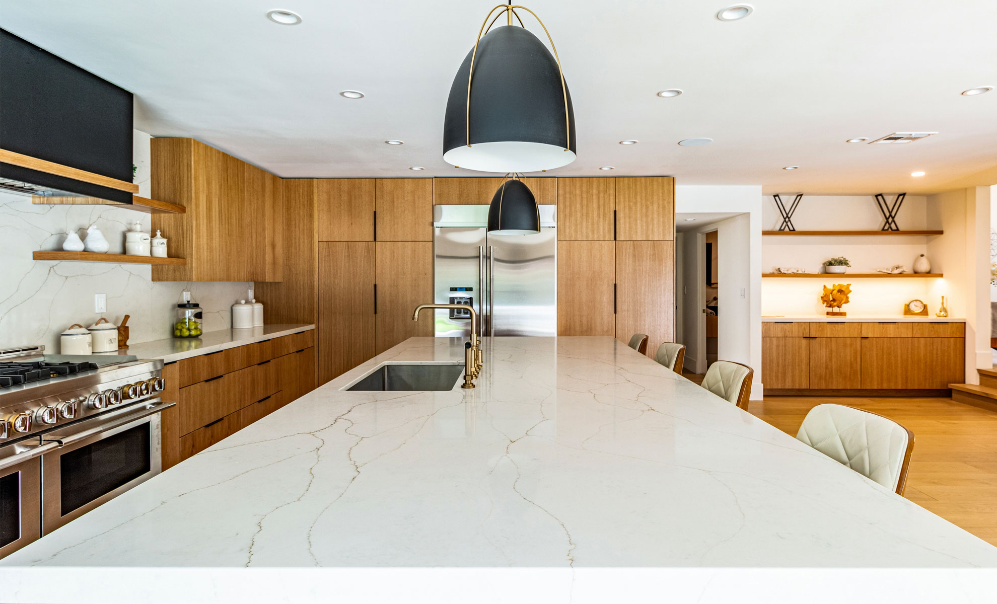 This kitchen features wooden cabinets, a white quartz countertop with veins, stainless steel appliances and two hanging light fixtures. There are white dishes displayed on the counters and on floating shelves.
